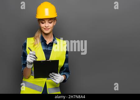 Costruttore sorridente in hardhat e gilet di sicurezza che tiene penna e appunti isolato su grigio Foto Stock