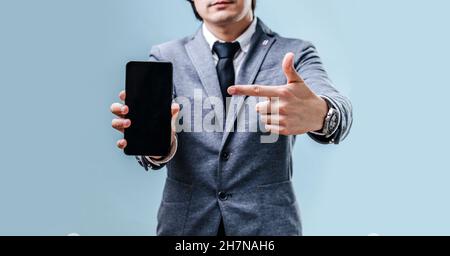 Una giovane bruna uomo d'affari in una giacca, una camicia e una cravatta tiene il telefono e punta il dito verso il telefono. Primo piano dello schermo vuoto sul retro blu Foto Stock