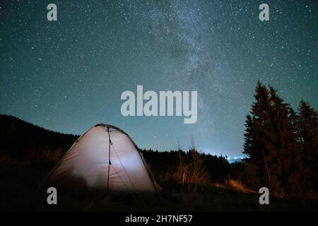 Luminosa tenda turistica illuminata che risplenderà sul campeggio in montagne scure sotto il cielo notturno con stelle scintillanti. Concetto di stile di vita attivo. Foto Stock