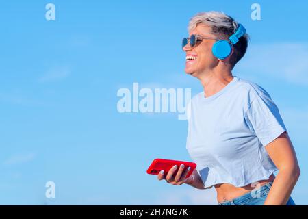 bella giovane donna con capelli biondi corti, occhiali da sole e breve t-shirt che mostra il suo pulsante della pancia ha divertimento e sorride ascoltando musica con wireless lui Foto Stock
