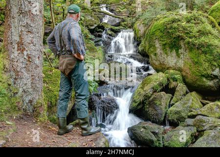 Un zaino in spalla si erge di fronte ad una piccola cascata e gode il suono dell'acqua e del silenzio nella foresta. Foto Stock