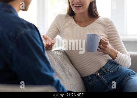 Giovane moglie europea allegra con una tazza che parla con il marito in un accogliente soggiorno interno Foto Stock