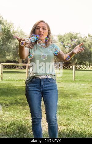 Le donne che indossano t-shirt e jeans che giocano con bolle di sapone in un parco, tre quarti di colpo donna caucasica di mezza età che si divertono ioutdoor in un'estate. Ma Foto Stock