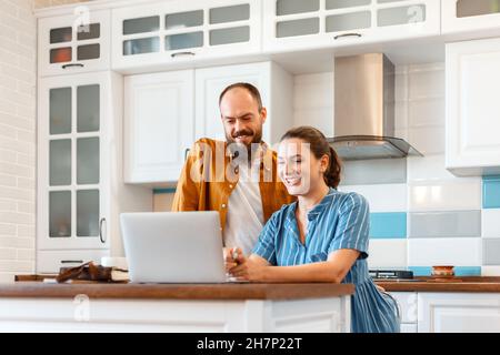 Sorridente giovane coppia sposata che parla tramite videochiamata utilizzando un computer portatile a casa in cucina. Donna e uomo felici di sentire buone notizie da e-mail in Foto Stock