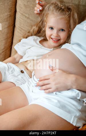 La figlia felice mostra il ventre come il ventre incinta della mamma per divertimento. Ritratto di bambino sorridente e madre incinta. Maternità amore relazione Foto Stock