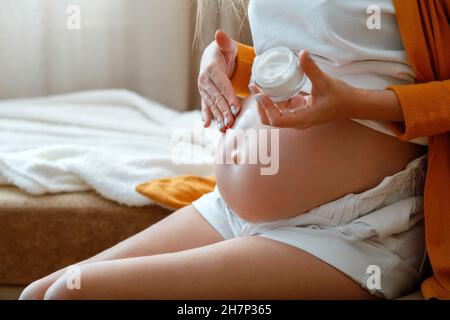 La donna incinta le spalma il ventre con crema di lozione a casa. Assistenza sanitaria durante la maternità e la gravidanza. Cura quotidiana della pelle del ventre incinta. Irriconoscibile Foto Stock