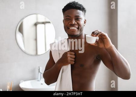 Black Guy che mostra crema idratante vaso a macchina fotografica in bagno Foto Stock