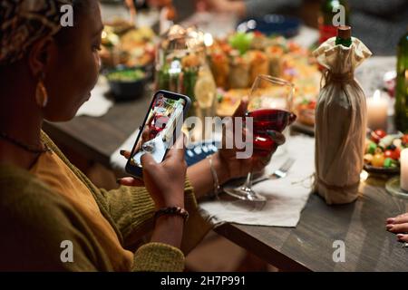 Giovane donna africana che fa foto di vino rosso sul suo telefono cellulare mentre si siede al tavolo da pranzo Foto Stock