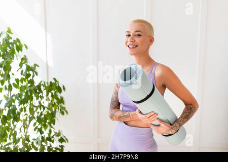 Ritratto di donna positiva che tiene il tappeto yoga e sorride alla macchina fotografica mentre si alza contro la parete bianca, pronto per l'allenamento Foto Stock