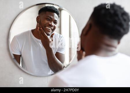 Nero maschio toccare unshaven Chin vicino specchio in bagno moderno Foto Stock