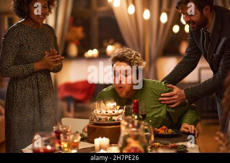 Giovane seduto al tavolo e che soffia le candele sulla torta di compleanno insieme ai suoi amici Foto Stock