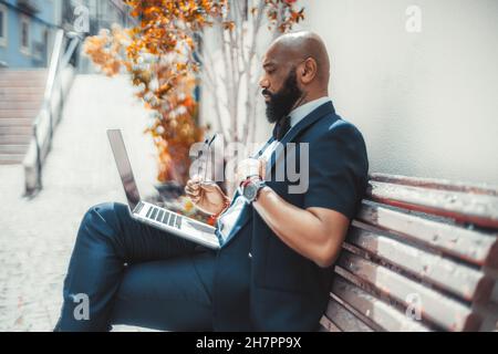 Vista laterale di un serio calvo maturo elegante uomo d'affari nero con una barba ben curata, utilizzando il suo computer portatile mentre si siede su una panca di strada in legno con e. Foto Stock