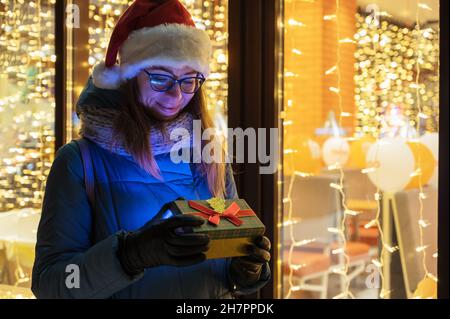 Graziosa Donna in cappello Santas con scatola di regali vicino alla finestra del caffè illuminato. Lei ondeggia la mano e attende il suo amato compagno. Natale presenta vacanze, Foto Stock