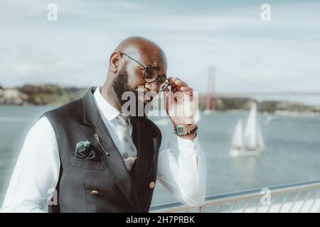 Un cappero afroamericano senior con una barba nera ben curata e in un abito alla moda formale con un gilet sta regolando il suo elegante occhiale whil Foto Stock