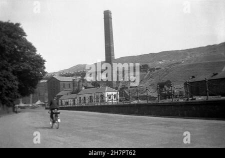 1935, storico, un giovane che pedala oltre una collisione nella valle a Ebbw vale, Galles, Regno Unito. Dal the1800s, Ebbw vale si era sviluppata in un importante centro per la produzione di ferro e acciaio, alimentato dal carbone proveniente dalle numerose miniere della zona. Nella foto si può vedere la miniera stack o camino che è stato attaccato al motore e che è interessante una costruzione quadrata in contrasto con il normale round. Foto Stock
