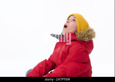 Primo piano di un bel ragazzo europeo di età elementare sorpreso stupito in giallo chiaro cappello di lana e parka rossa guardando in su con bocca aperta contro bianco Foto Stock