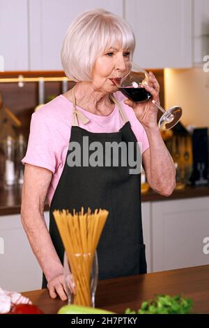la donna allegra senior beve vino rosso durante la cottura nella cucina moderna. Cibo, istruzione, concetto di stile di vita Foto Stock