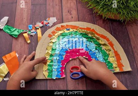 Le mani del bambino fanno un arcobaleno con carta colorata e forbici su un tavolo di legno Foto Stock