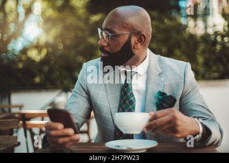 Un ritratto di un bel balsamo dapper maturo uomo nero imprenditore con una barba ben curata e in un abito formale con una cravatta, seduto in una strada Foto Stock