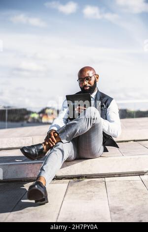 Un colpo verticale di un uomo d'affari elegante e nero calvo per adulti con una barba ordinata, in occhiali e un abito alla moda con un gilet, utilizzando un futu incandescente Foto Stock