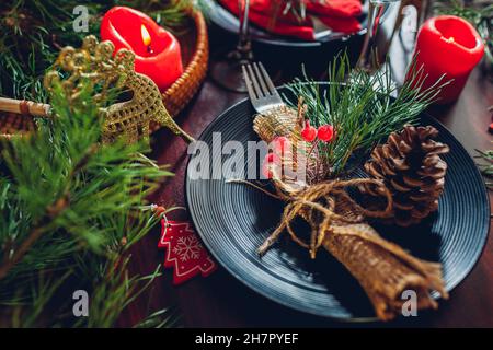 Cena natalizia con tavolo per due, decorato con rami di abete, candele rosse, coni, ornamenti. Piatti con posate e bicchieri circondati Foto Stock