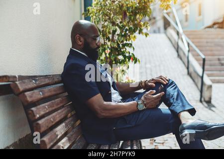 Una vista laterale di un imprenditore africano-americano maturo balvo con una barba nera ben curata, in occhiali e abito formale blu, seduto su un s Foto Stock