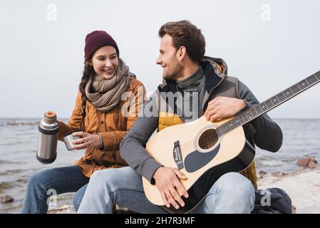 donna allegra con thermos e uomo con chitarra seduto sul lungofiume durante la passeggiata autunnale Foto Stock
