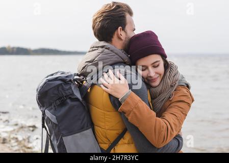 donna felice con occhi chiusi abbracciando il ragazzo con zaino mentre cammina vicino al fiume Foto Stock