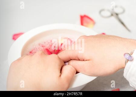 Le mani in ciotola bianca bagnano le unghie con acqua e petali di rosa per ammorbidire la cuticola e la pelle asciutta Foto Stock