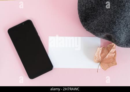 Smartphone, un biglietto d'auguri vuoto, un cappello e una foglia d'autunno asciutta su una scrivania rosa Foto Stock