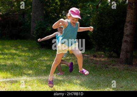 Due bambini attivi e gioiosi, ragazze che giocano con l'acqua nel cortile, saltando, correre intorno, sorelle fratelli divertimento estate, acqua tubo, divertirsi Foto Stock