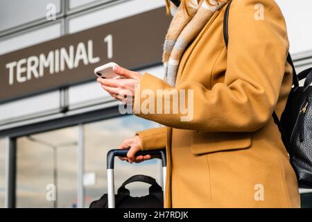 Donna turistica in colorato cappotto con sciarpa legge messaggio su smartphone tenendo maniglia valigia presso il terminal internazionale aeroporto vicino vista Foto Stock