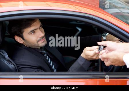 Un giovane uomo d'affari in una tuta nera che maneggia la chiave dell'auto al personale addetto al servizio di assistenza mentre tiene la ruota dell'auto. Foto Stock