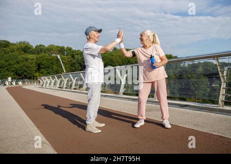 L'uomo anziano e la donna gioiosi in abbigliamento sportivo si allenano insieme su un ponte pedonale Foto Stock
