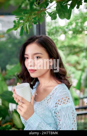 Appendere le piante in vaso in estate con la donna in piedi sul portico di casa nella mattina di cabina in legno cottage bere il tè o caffè Foto Stock