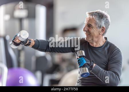 Uomo anziano che si allenava con i manubri all'interno della palestra, sorridendo. Foto Stock