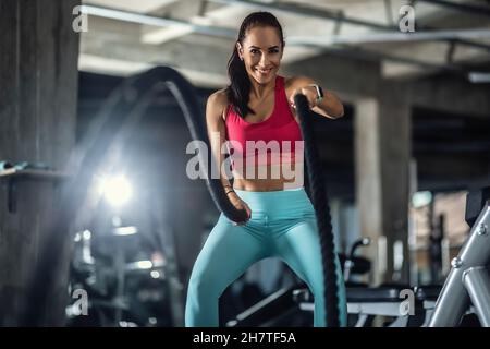 La donna che usa le corde del crossfit funziona fuori in palestra, sorridendo. Foto Stock