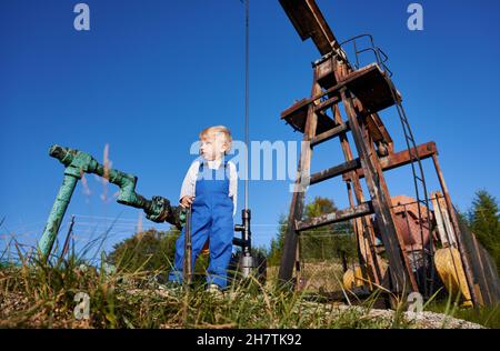 Vista ad angolo basso del bambino che si trova sullo sfondo della pompa oscillante dell'olio. Piccolo ragazzo in piedi sotto il cielo blu aperto, tenendo in una chiave a mano. Foto Stock