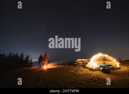 Giovani uomini e donne viaggiatori che tengono le mani mentre si levano in piedi vicino falò sotto il cielo notturno con le stelle. Splendida vista sul cielo stellato notturno sulla collina erbosa. Concetto di escursioni, campeggio notturno, relazioni Foto Stock