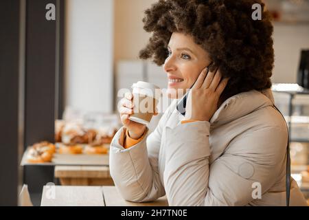 Donna felice 40 anni al caffè bere caffè e parlare telefono cellulare, acconciatura afro, godendo momento, persone felici, vita di città Foto Stock