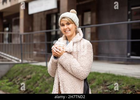 Giovane donna graziosa, di 28 anni, con cappello a maglia, cappotto in pelliccia di finta beige, che si riscalda il giorno d'inverno e che tiene una tazza riciclabile di caffè o tè, Foto Stock