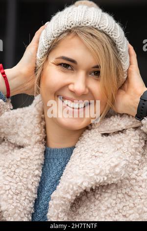 Ritratto di giovane, donna sicura, sorridente, guardando la macchina fotografica, indossando cappello a maglia e cappotto in pelliccia sintetica. Stile Street. Capelli biondi, bangs, occhi marroni. Foto Stock