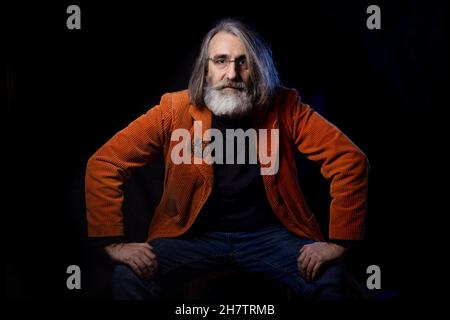 Аn intellettuale in occhiali alla moda guarda la fotocamera con uno sguardo critico. Un uomo con bacchette dai capelli grigi in giacca arancione. Foto Stock