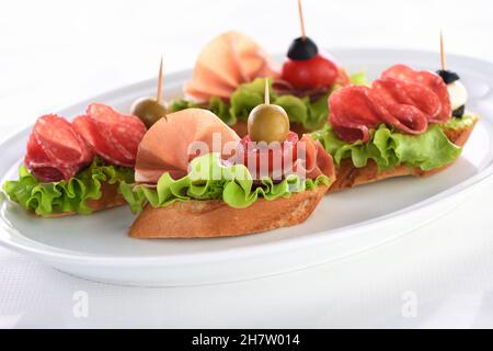 Tartine di baguette tenue con lattuga, salame o prosciutto di Parma, pomodori, mozzarella e olive. Piatto assortito di prelibatezze per la festa. Foto Stock