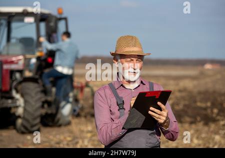 L'agricoltore senior scrive note in blocco note sul campo con il trattore in background Foto Stock
