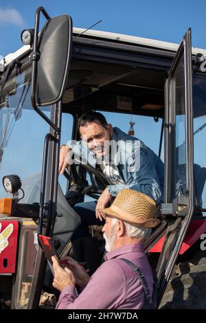 Conducente maturo seduto sul trattore e che parla con l'agricoltore senior accanto al veicolo sul campo Foto Stock