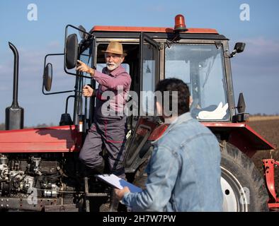 Due agricoltori che parlano sul campo. Uomo anziano in piedi sul trattore e che mostra qualcosa per adulti maturi sul campo Foto Stock