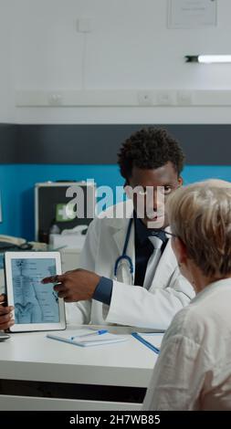 Giovane fisioterapista che mostra compresse di lesioni alle ossa per pazienti anziani con osteoporosi alla scrivania in ufficio medico. Donna anziana che guarda le vertebre virtuali per un esame medico Foto Stock