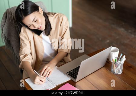 Scatto ad angolo superiore di donna asiatica che lavora con il laptop, risponde alle chiamate telefoniche e scrive in taccuino. La ragazza prende appunti mentre parla sullo smartphone Foto Stock