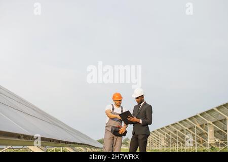 Uomo afro-americano in abito nero e casco bianco in piedi con tecnico indiano in uniforme su fattoria solare. Persone multirazziali che esaminano la produzione di energia verde pulita. Foto Stock